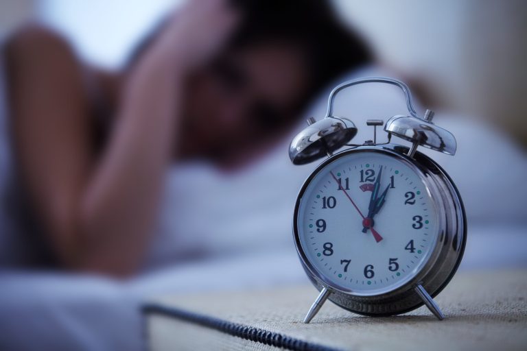 Réveil mécanique au premier plan avec une personne allongée, floue en arrière-plan, semblant ne pas dormir.